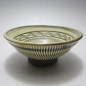 May include: A ceramic bowl with a cream-colored interior and a dark brown exterior. The bowl features a decorative pattern of vertical lines and wavy lines. The bowl is round and sits on a dark brown base.