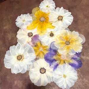 May include: A collection of translucent, layered paper flowers in shades of white, yellow, and purple. Each flower features delicate, thin petals with a textured appearance and a dark, circular center, arranged in a cluster with overlapping edges.