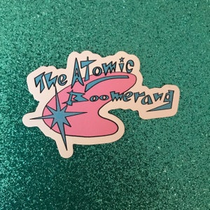 May include: A white sticker with a pink boomerang shape and a blue star. The text "The Atomic Boomerang" is written in blue.