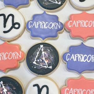 May include: A collection of twelve round and scalloped sugar cookies decorated with royal icing. Designs include the word Capricorn in white, the black Capricorn zodiac symbol on white, and a marbled black space background with white constellations.