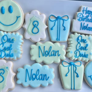 May include: A collection of decorated sugar cookies in shades of light blue and pale green. Designs include smiley faces, gift boxes, tiered cakes with the text Happy Birthday Nolan, and scalloped shapes featuring the text One Cool Dude or Nolan.