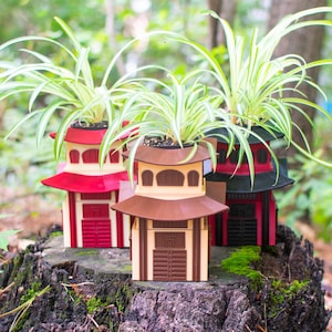 Può includere: Tre fioriere a forma di pagoda, ognuna con una pianta ragno. Le fioriere sono nei toni del rosso, marrone e beige, con un design architettonico giapponese. Le fioriere sono su un ceppo d'albero.