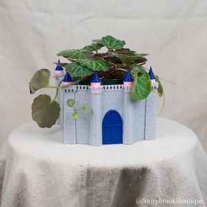Puede incluir: Una maceta gris con forma de castillo con una puerta azul y torretas rosas y azules, que contiene una planta verde con hojas estampadas. La maceta está sobre una superficie blanca.