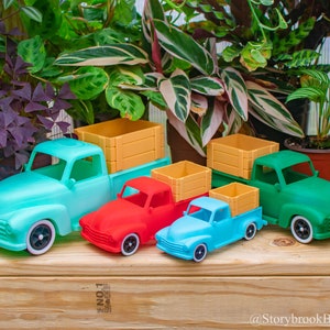 Puede incluir: Una colección de camiones de juguete coloridos en varios tamaños y tonos, incluyendo turquesa, rojo, azul y verde. Cada camión tiene una caja de estilo madera. Los camiones están colocados sobre una superficie de madera, con exuberantes plantas verdes en el fondo.