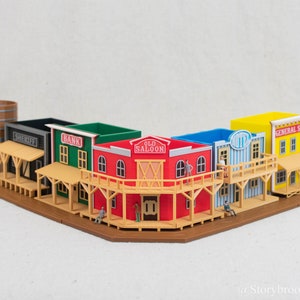 Może przedstawiać: Miniaturowa diorama miasteczka na Dzikim Zachodzie z budynkami z szyldami banku, szeryfa i sklepu ogólnego. Budynki są czerwone, zielone, niebieskie i żółte z drewnianymi akcentami. Zawiera również wieżę ciśnień i miniaturowe figurki.