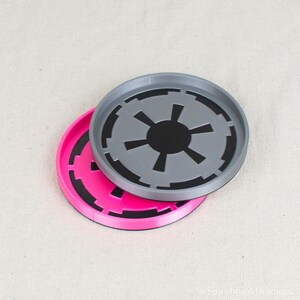Puede incluir: Dos posavasos redondos, uno rosa intenso y otro gris, cada uno con un diseño negro de estrella estilizada. Los posavasos están hechos de un material duradero y están diseñados para proteger las superficies de las marcas de bebidas.