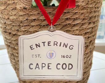 May include: A clear acrylic sign with the words "ENTERING CAPE COD" in black lettering, with "EST. 1602" above. The sign is suspended by a red velvet ribbon from a woven basket filled with greenery and white flowers.