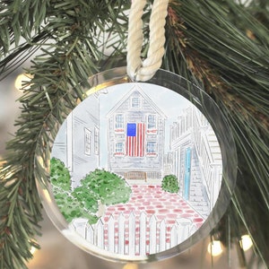 May include: A round, clear acrylic Christmas ornament featuring a watercolor illustration of a house with an American flag hanging on the side. The house has a red brick patio and a white picket fence.