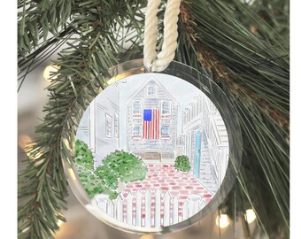 May include: A round, clear acrylic Christmas ornament featuring a watercolor illustration of a house with an American flag hanging on the side. The house has a red brick patio and a white picket fence.