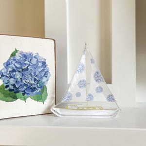 Pode incluir: Um veleiro de vidro transparente com padrões florais azuis e detalhes dourados está numa prateleira branca. À esquerda, um bloco de madeira com uma impressão de hortênsia azul. Objeto decorativo náutico.