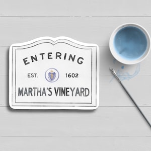 May include: A white sign with black text that reads "Entering Est. 1602 Martha's Vineyard". The sign has a rounded top and a small blue and gold seal in the center. A blue paint dish and a gray paintbrush are on a white wooden surface.