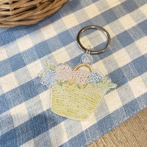 May include: A white wicker basket filled with pink and blue flowers, attached to a silver key ring. The basket is a charm for a keychain.