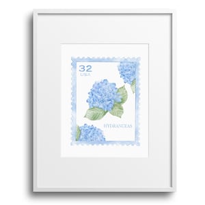 Op de afbeelding: Een ingelijste print met een aquarelillustratie van blauwe hortensia's op een postzegelontwerp. De postzegel leest "32 USA" en "HYDRANGEAS" in een lichtblauw kader. De print zit in een witte lijst.