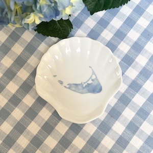 May include: A white ceramic dish shaped like a seashell with a blue outline of the island of Martha's Vineyard. The dish is on a blue and white gingham tablecloth.