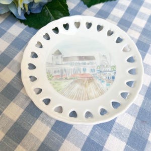 Peut inclure: Assiette en céramique blanche avec un motif découpé en forme de cœur autour du bord. Le centre de l'assiette présente une illustration aquarelle d'un bâtiment avec une promenade en bois devant.