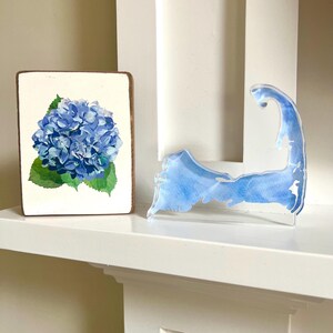 Pode incluir: Um bloco de madeira com uma pintura em aquarela de uma flor de hortênsia azul e folhas verdes. Ao lado, uma forma clara de acrílico de Cape Cod com detalhes em aquarela azul. Ambos os itens são exibidos em uma prateleira branca.