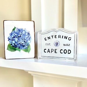 Pode incluir: Duas placas decorativas numa prateleira branca. Uma é uma placa retangular de madeira com uma pintura em aquarela de uma hortênsia azul. A outra é uma placa de acrílico transparente que diz "Entering Cape Cod" com a data de fundação de 1602.