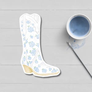 May include: A white cowboy boot sticker with blue floral accents and a light yellow heel. A small white bowl with blue liquid and a paintbrush are next to the boot on a light gray surface.