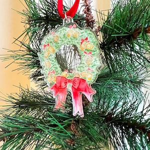 May include: A green Christmas wreath ornament with a red ribbon and gold bells. The ornament is made of clear plastic and has a vintage look.