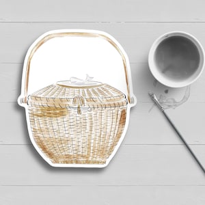 May include: A brown wicker basket with a lid and a white handle. The basket is sitting on a white wooden surface.