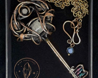 May include: A handcrafted wire jewelry necklace featuring a key pendant with a gemstone and bead embellishments. The key is made of brass and copper wire, with a chain and a black box. The pendant has a gemstone and bead embellishments.