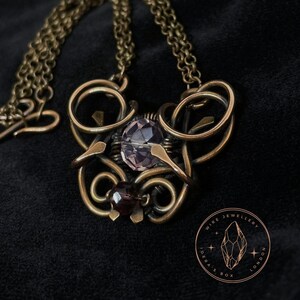 May include: A delicate bronze wire necklace with a purple gemstone and a smaller red gemstone. The necklace is on a black velvet background. The pendant is in the shape of a swirl with a small red gemstone at the bottom. The necklace is from Wire Ideas Box Jewellery London.