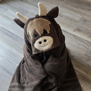 Op de afbeelding: Een bruine handdoek met capuchon in de vorm van een Schotse hooglander. De handdoek heeft een lichtbruin gezicht met een lachende mond, zwarte ogen en beige hoorns. De handdoek is opgevouwen en ziet er zacht en pluche uit.