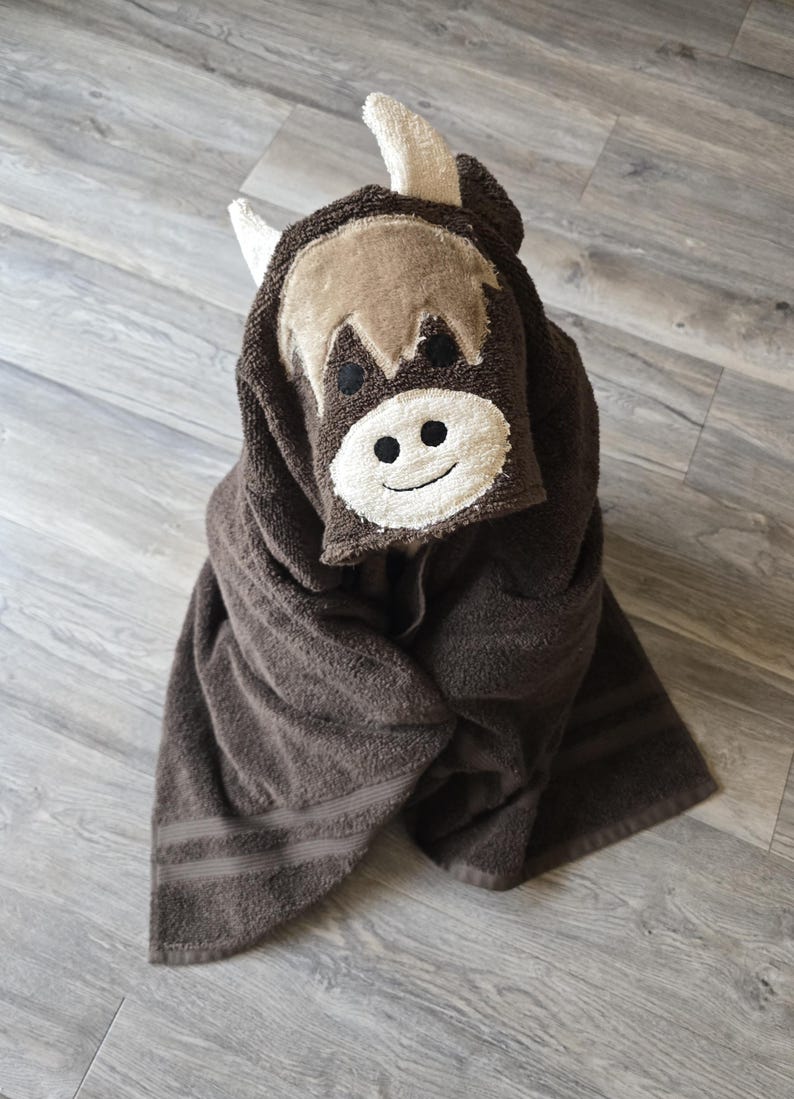 Op de afbeelding: Een bruine badhanddoek met capuchon en een Highland-koeienontwerp. De capuchon heeft een lachend gezicht met witte hoorns en een lichtbruine franje. De handdoek is opgevouwen en toont het zachte, absorberende materiaal.