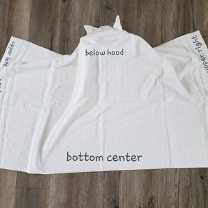 Op de afbeelding: Witte badhanddoek met capuchon en een getextureerd oppervlak. De handdoek heeft een capuchon met puntige oren. De woorden "upper left", "center left", "lower left", "bottom center", "below hood", "lower right", "center right" en "upper right" staan op de handdoek.