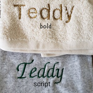 Op de afbeelding: Twee handdoeken, &eacute;&eacute;n cr&egrave;mekleurig en &eacute;&eacute;n grijs, met de naam "Teddy" erop geborduurd. De cr&egrave;mekleurige handdoek heeft "Teddy" in gouden, vette letters en de grijze handdoek heeft "Teddy" in groen script.