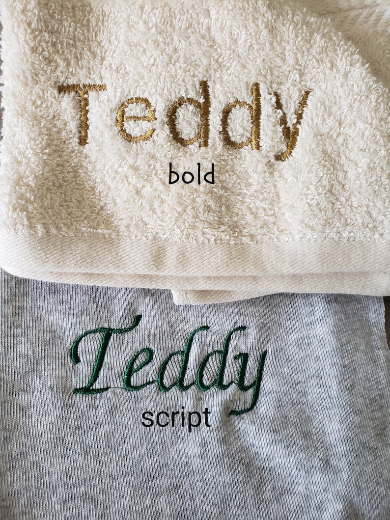 Op de afbeelding: Twee handdoeken, &eacute;&eacute;n cr&egrave;mekleurig en &eacute;&eacute;n grijs, met de naam "Teddy" erop geborduurd. De cr&egrave;mekleurige handdoek heeft "Teddy" in gouden, vette letters en de grijze handdoek heeft "Teddy" in groen script.