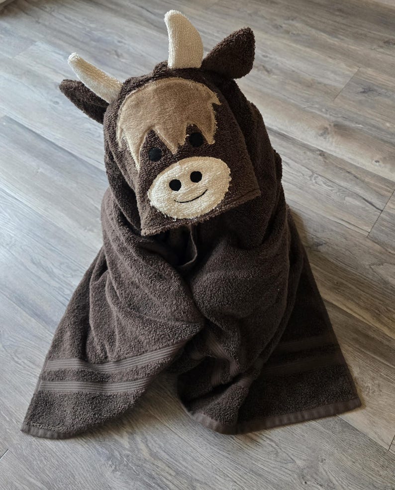 Op de afbeelding: Een bruine handdoek met capuchon in de vorm van een Schotse hooglander. De handdoek heeft een lichtbruin gezicht met een lachende mond, zwarte ogen en beige hoorns. De handdoek is opgevouwen en ziet er zacht en pluche uit.