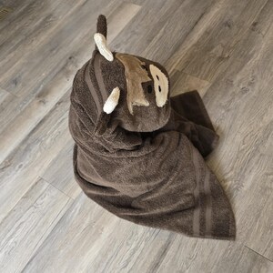 Op de afbeelding: Een bruine badhanddoek met capuchon, gevouwen in de vorm van een cartoon dier. De handdoek heeft een lichtbruin gezicht met witte accenten en een paar witte hoorns. De handdoek is gevouwen op een lichtgrijze houten vloer.