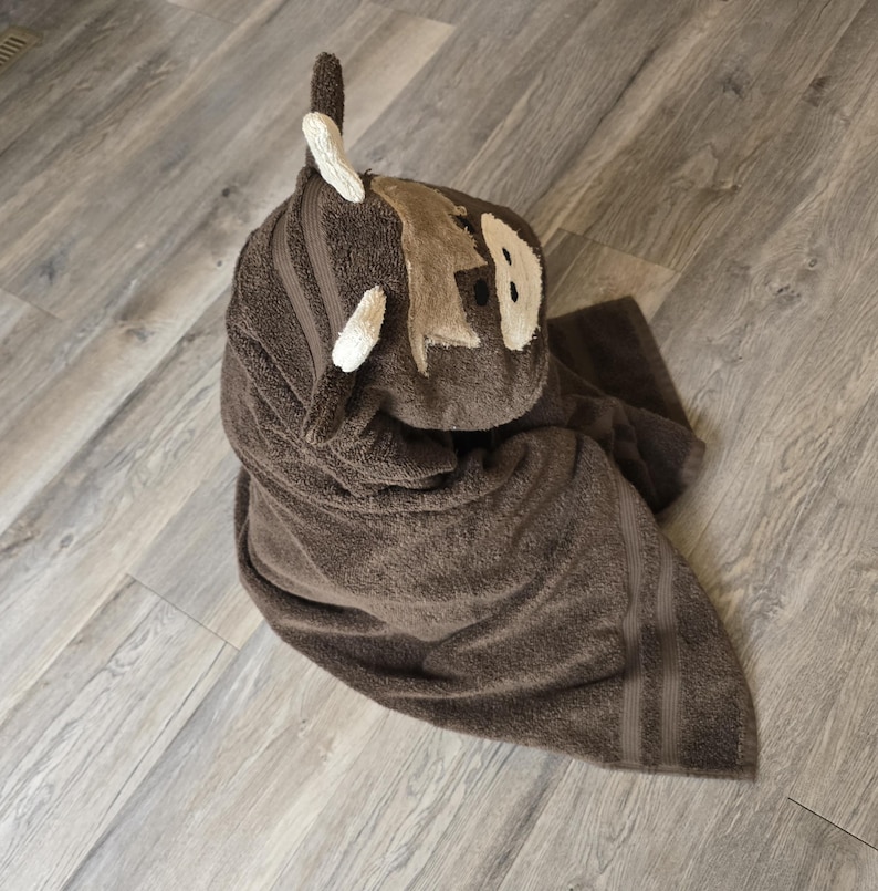 Op de afbeelding: Een bruine badhanddoek met capuchon, gevouwen in de vorm van een cartoon dier. De handdoek heeft een lichtbruin gezicht met witte accenten en een paar witte hoorns. De handdoek is gevouwen op een lichtgrijze houten vloer.