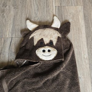 Op de afbeelding: Een bruine badhanddoek met capuchon en een koeiengezicht. De capuchon heeft witte hoorns, een beige franje, zwarte ogen en een lachend gezicht. De handdoek is gemaakt van zacht, absorberend materiaal.