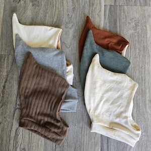 May include: A collection of six pointed pixie-style bonnets in various textures and colors, including cream, heather gray, dark brown, rust orange, and sage green. Each bonnet features a distinct elongated peak and a soft, folded fabric construction.