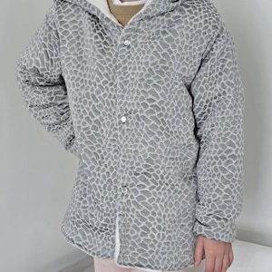 May include: A gray hooded jacket with a textured pattern and white lining. The jacket has a button-up front and a hood with a white interior. The jacket is worn over a light brown shirt and pink pants.