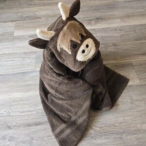 Op de afbeelding: Een bruine handdoek met capuchon in de vorm van een Schotse Hooglandkoe. De handdoek heeft een geborduurd gezicht met een lachende uitdrukking, kleine hoorns en een zachte, pluche textuur. De handdoek is opgevouwen en toont zijn absorberende materiaal.