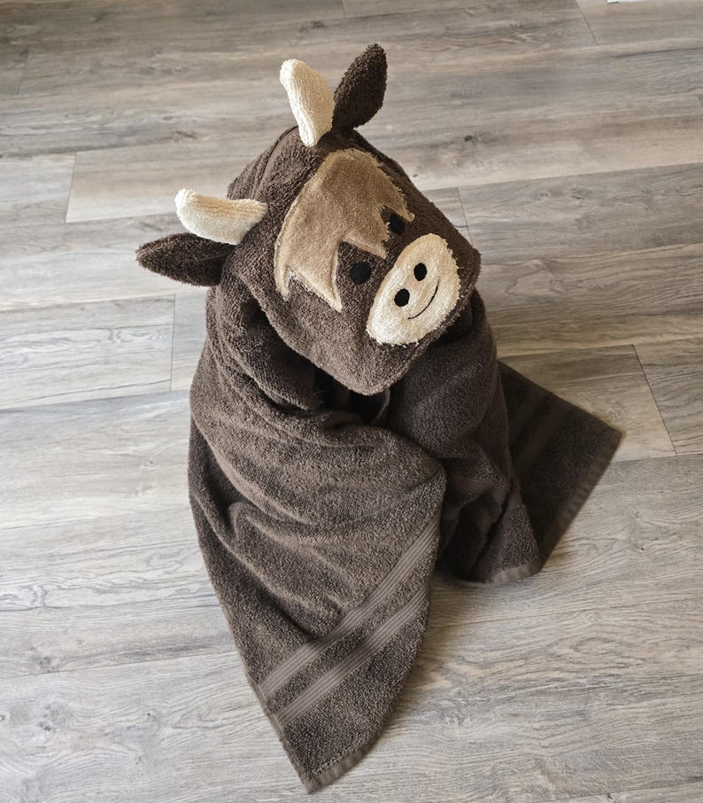 Op de afbeelding: Een bruine handdoek met capuchon in de vorm van een Schotse Hooglandkoe. De handdoek heeft een geborduurd gezicht met een lachende uitdrukking, kleine hoorns en een zachte, pluche textuur. De handdoek is opgevouwen en toont zijn absorberende materiaal.