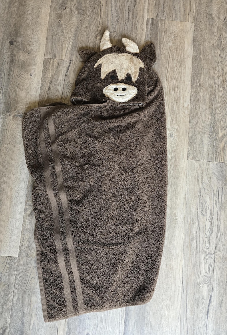 Op de afbeelding: Een bruine badhanddoek met capuchon en een Highland-koeienontwerp. De handdoek heeft een koeiengezicht op de capuchon met witte en beige accenten. Twee verticale strepen lopen langs de zijkant. De handdoek is gemaakt van zacht, absorberend materiaal.