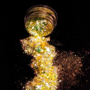 Puede incluir: Un tarro transparente con purpurina dorada, verde e iridiscente que se derrama sobre un fondo negro.