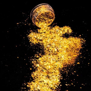 Puede incluir: Un frasco transparente con purpurina dorada que se derrama sobre un fondo negro. La purpurina está dispersa en forma de nube.