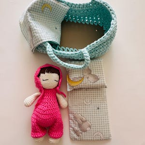May include: A crocheted doll wearing a pink outfit and a hooded jacket, lying on a blue and white checkered blanket with a pillow. The doll is in a blue crocheted basket with a handle.