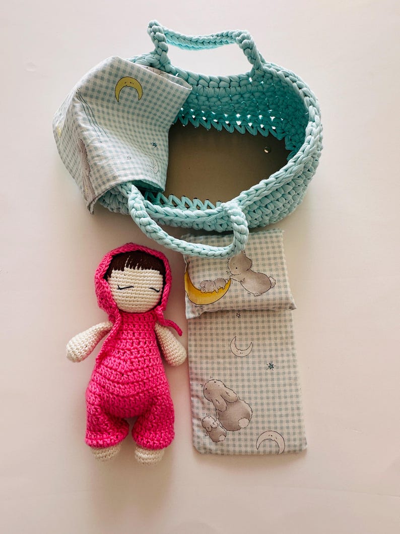 May include: A crocheted doll wearing a pink outfit and a hooded jacket, lying on a blue and white checkered blanket with a pillow. The doll is in a blue crocheted basket with a handle.