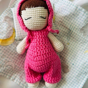 May include: A crocheted doll with pink clothing and a pink hooded hat, lying on a blue and white checkered blanket.