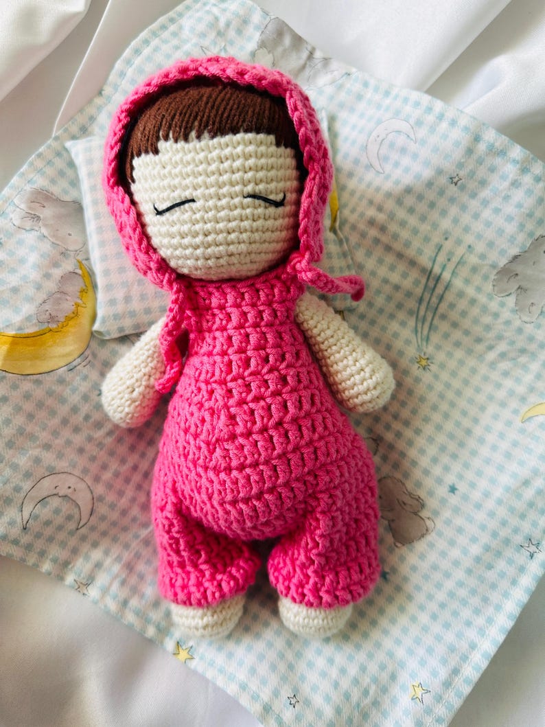 May include: A crocheted doll with pink clothing and a pink hooded hat, lying on a blue and white checkered blanket.