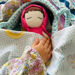 May include: A crocheted doll with brown hair and closed eyes, wearing a pink hooded dress, is tucked into a blue and white checkered blanket with a floral pattern. The doll is being held by a child's hand.