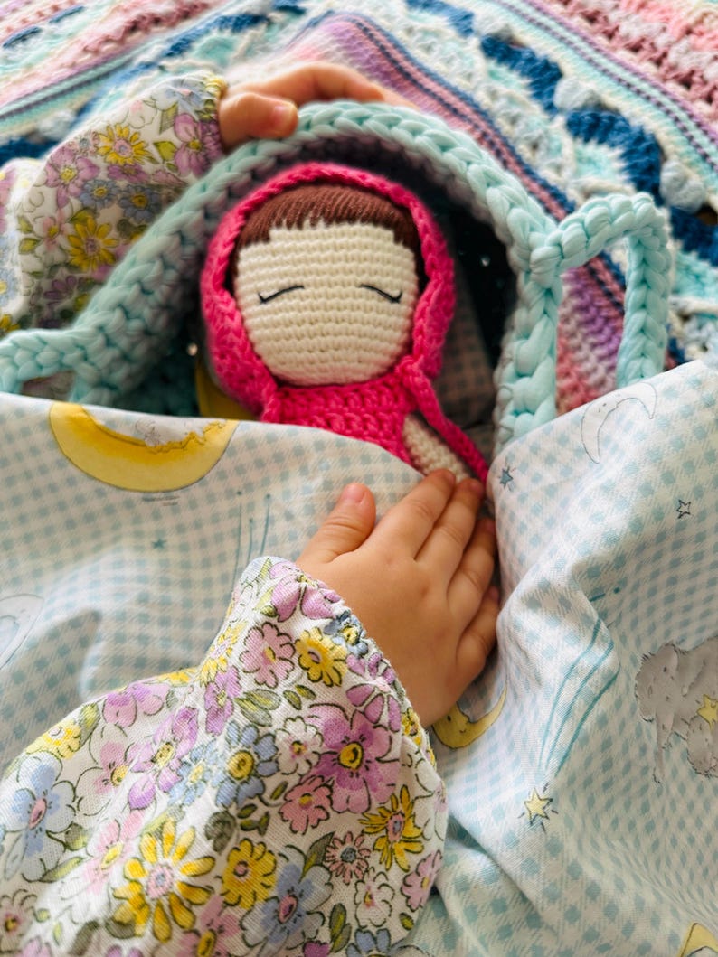 May include: A crocheted doll with brown hair and closed eyes, wearing a pink hooded dress, is tucked into a blue and white checkered blanket with a floral pattern. The doll is being held by a child's hand.