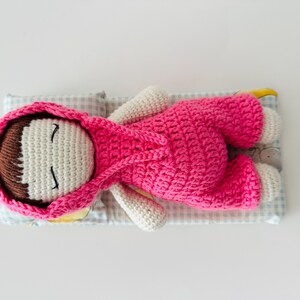 May include: A crocheted doll wearing a pink hooded outfit is lying on a blue and white checkered fabric. The doll has brown hair, white skin, and closed eyes.