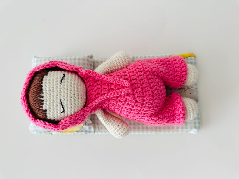 May include: A crocheted doll wearing a pink hooded outfit is lying on a blue and white checkered fabric. The doll has brown hair, white skin, and closed eyes.